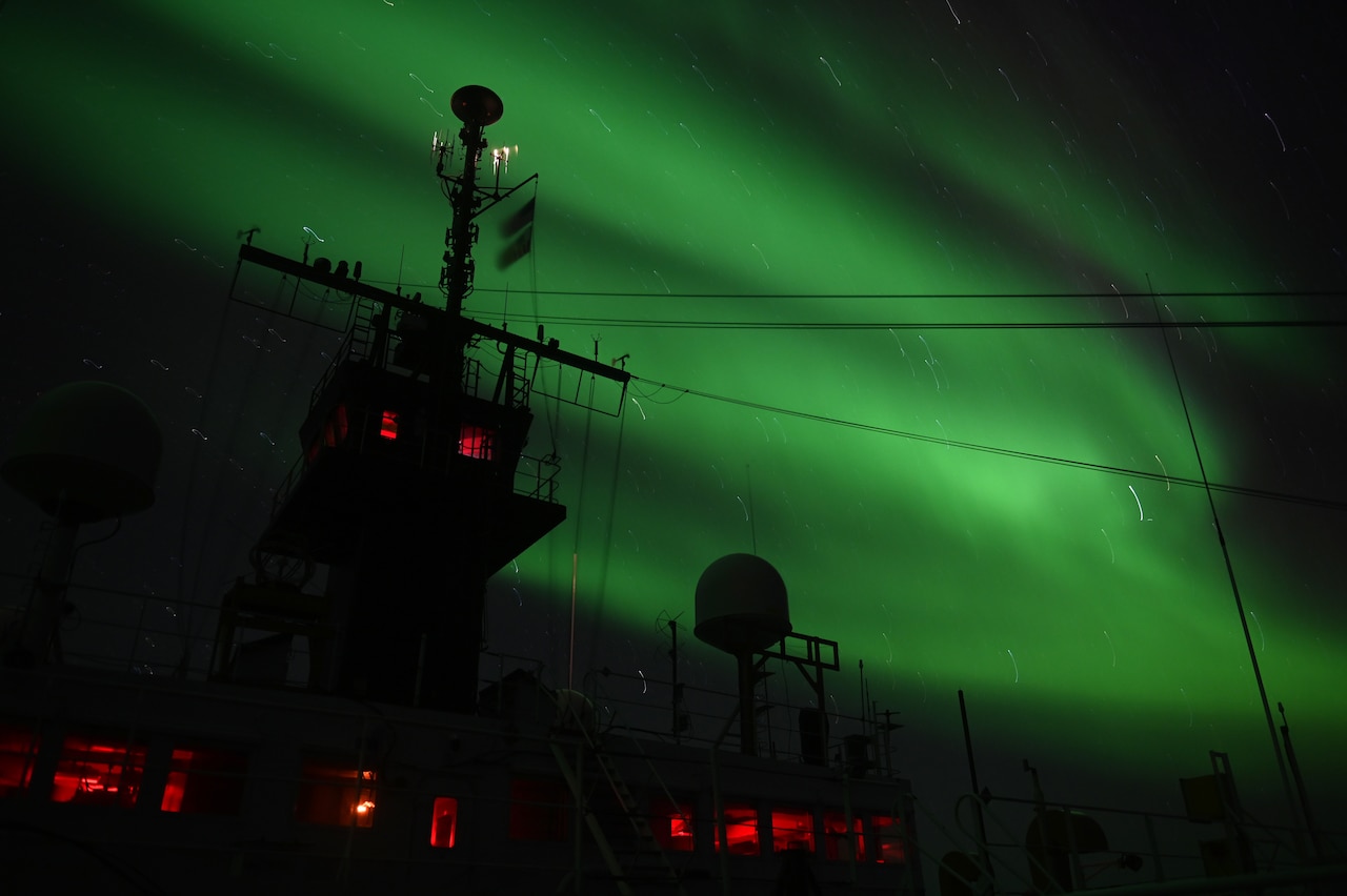 A green light in the sky backlights the cables of a ship in silhouette.