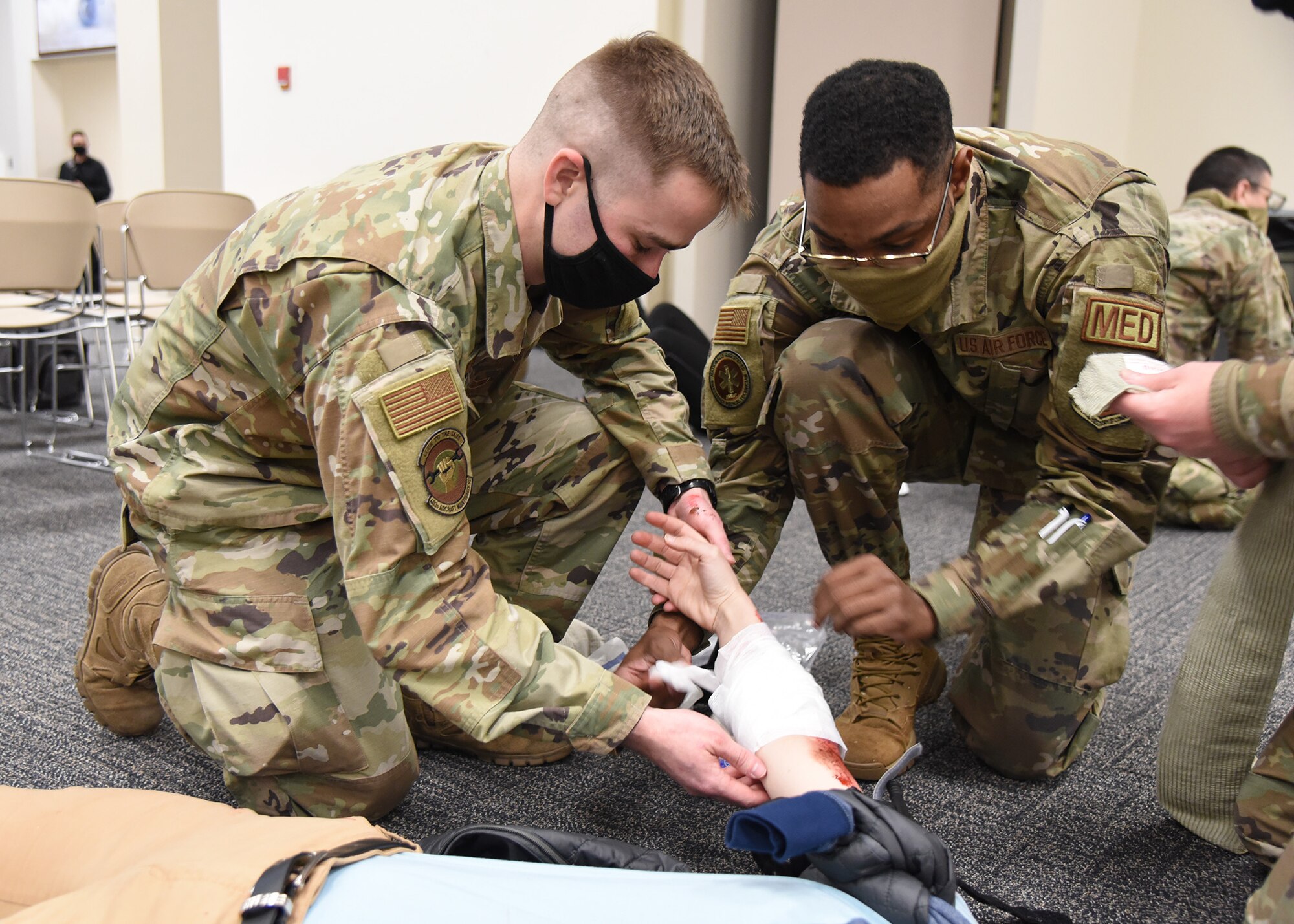Two Airmen kneel and wrap a person's arm with gauze.