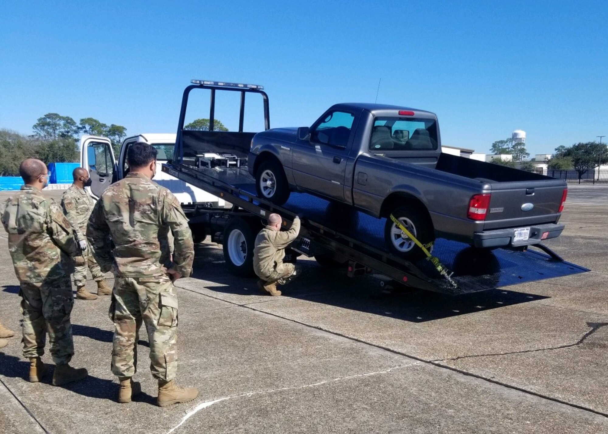 Three Airmen stand to the side and watch one fasten a pickup truck to tow