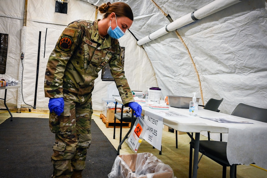 An airman wearing a face mask and gloves holds a plastic biohazard bag while placing it in a patient sample collection box.