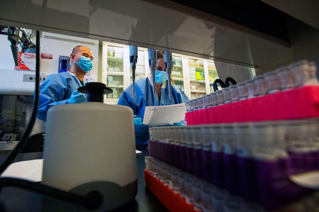 Two men wearing face masks and gloves review patient information before testing diagnostic samples.