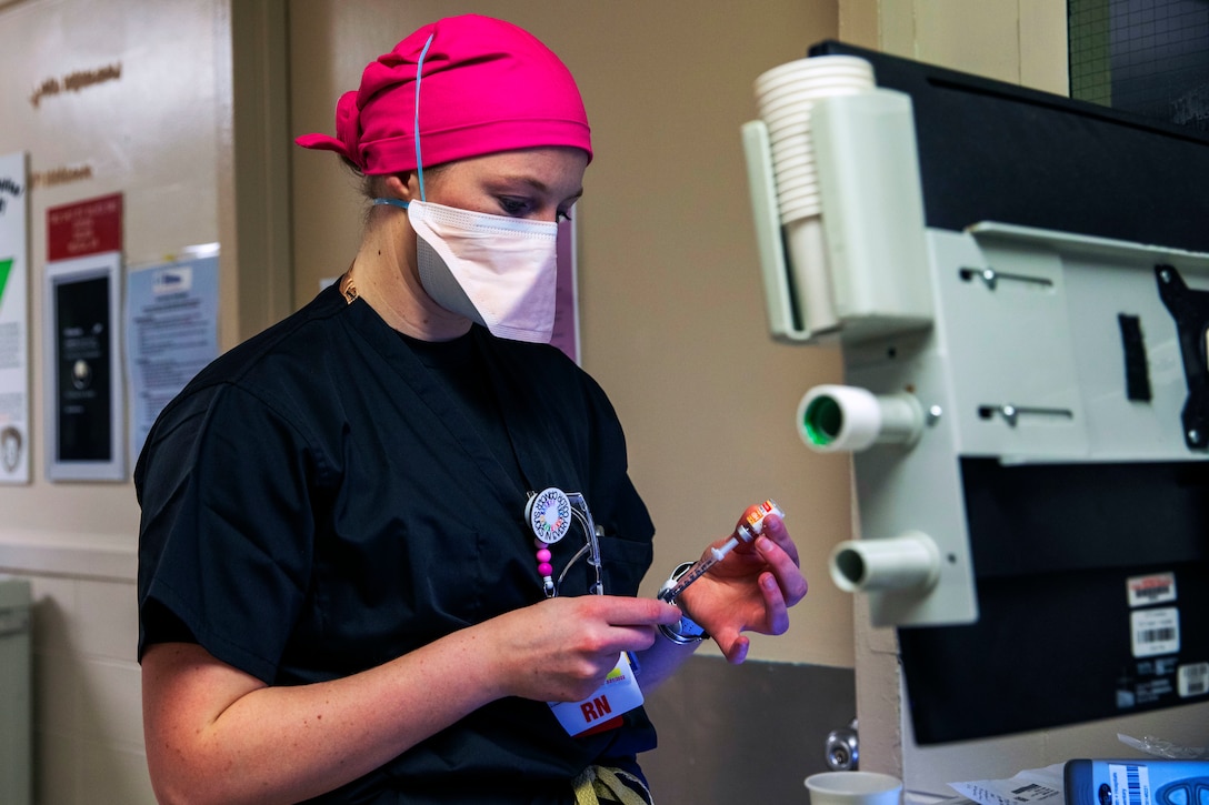 A soldier wearing a face mask holds a syringe that is inserted into a small bottle while drawing medication for a patient.