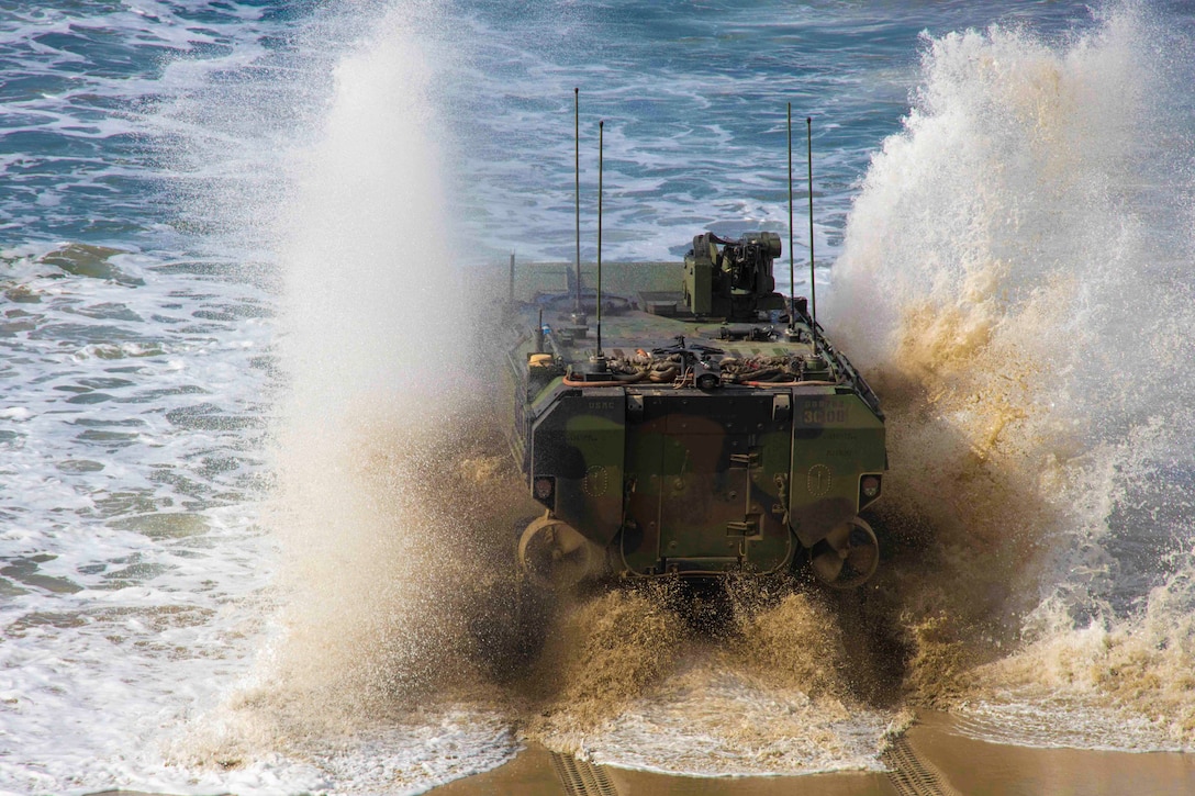 An amphibious combat vehicle drives on a beach as water sprays on either side.