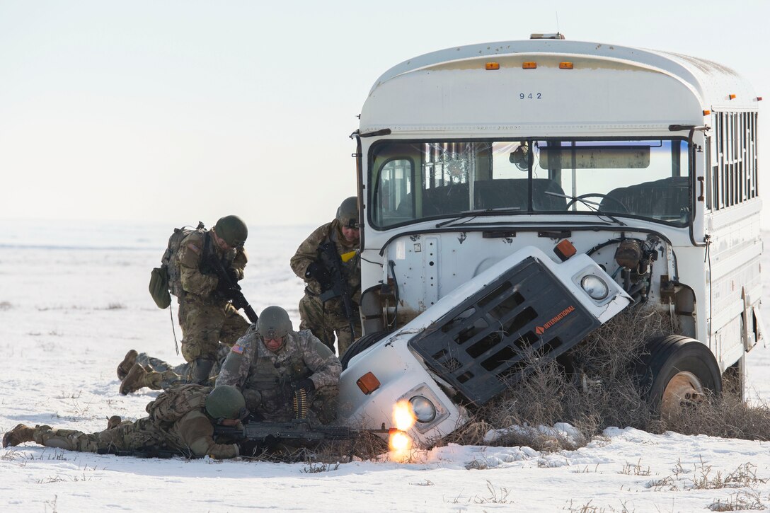 Soldiers, airmen, sailors and Marines holding weapons stand near a broken-down bus in a snowy field.