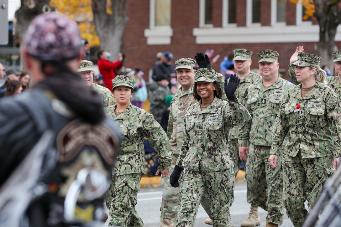 Service members walk down a street and wave to a crowd gathered on the sidewalk.