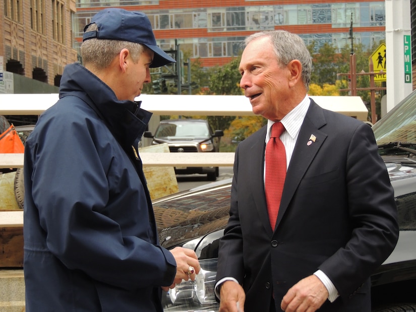 Two men stand outdoors and speak to each other.