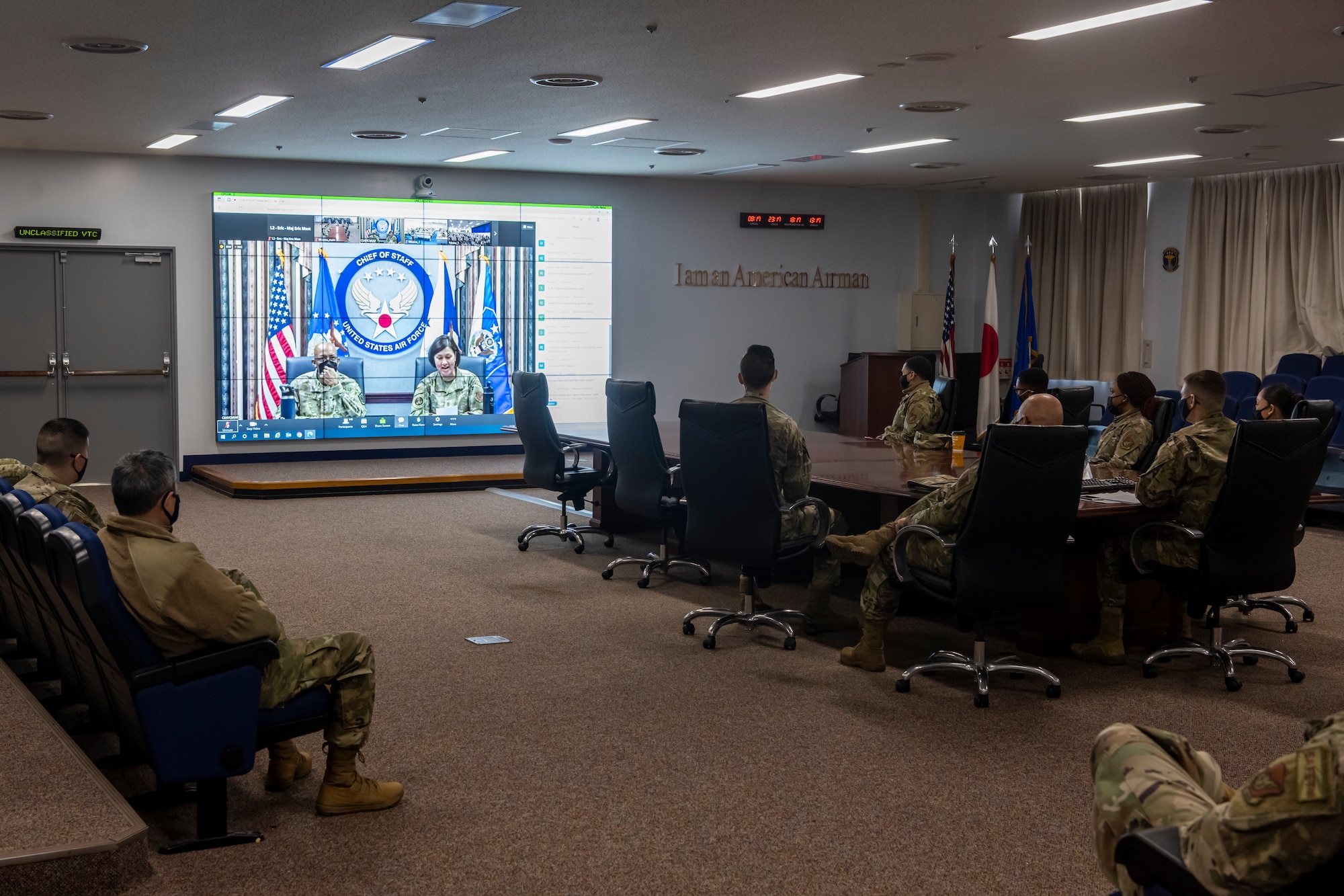 Airmen sitting in a conference room listen to a virtual zoom call.