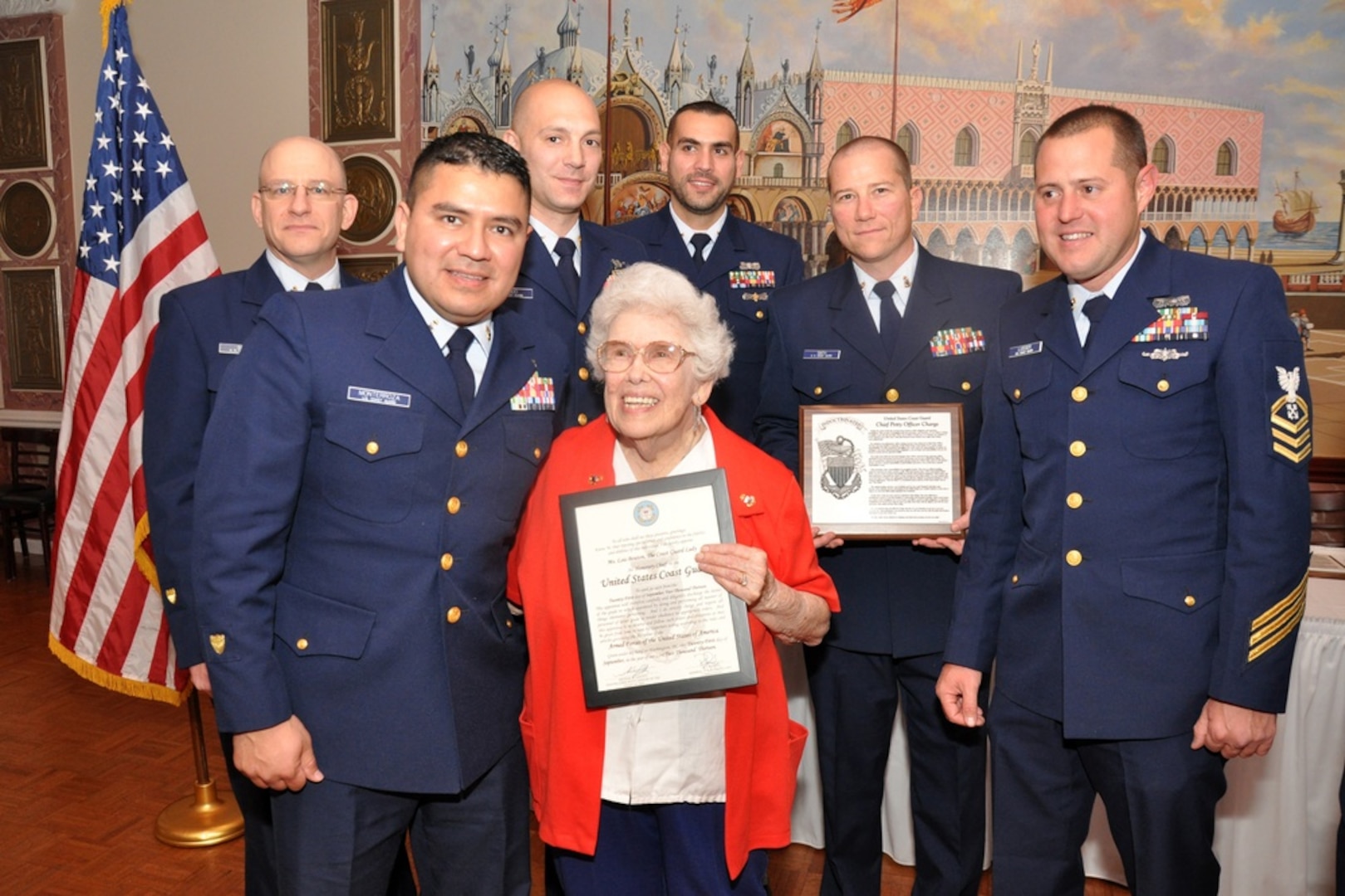 Coast Guard veteran in bright orange sweater stands in the center of uniformed Coast Guard members, receiving an award