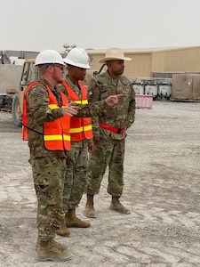 Three men in camo uniform orange safety vests and white hardhats stand outside while one points to the right.