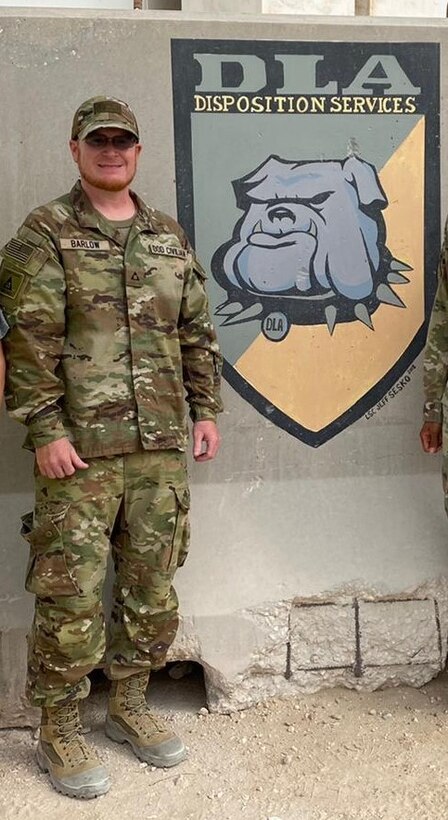 Man in a camo uniform stands against an outdoor wall with an emblem of a bulldog painted behind him.
