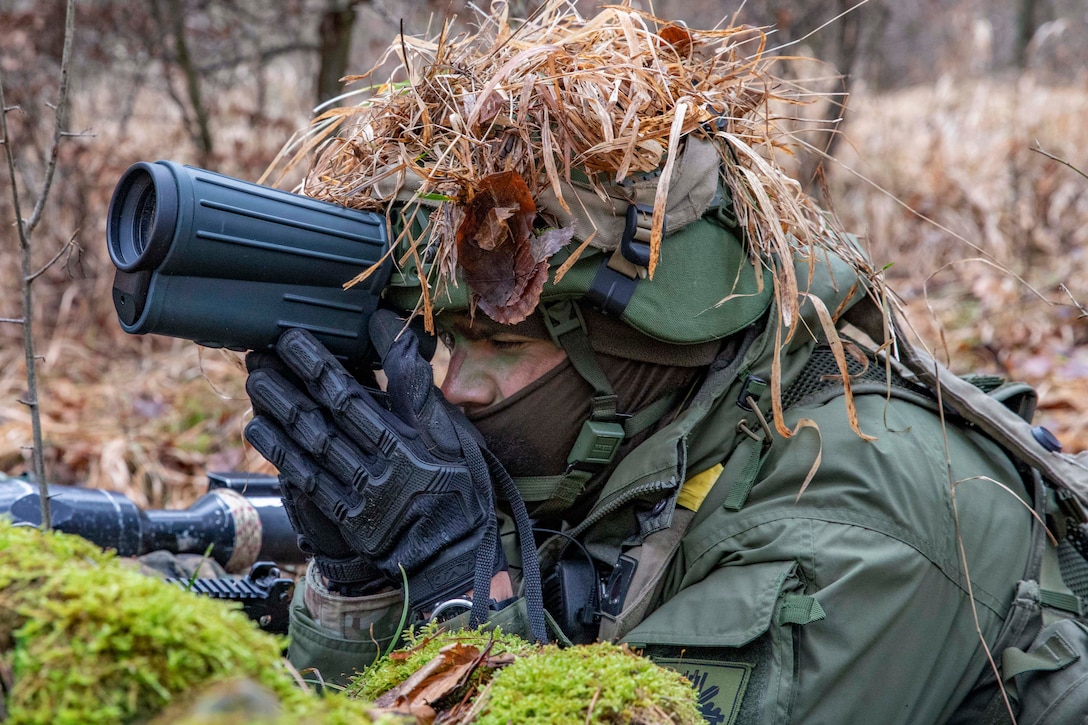 A soldier wearing camouflage uses binoculars while laying on the ground in a wooded area.