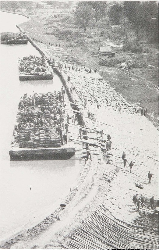 The Great Flood of 1927 created many unusual conditions that called for unusual remedies. Here we see a levee that is being paved on the landside slope, with sandbags overlaying wood and brush stabilizers. The levee has already been raised once, and is again in danger of being topped, thus it was necessary to reinforce the slope before attempting further repairs.