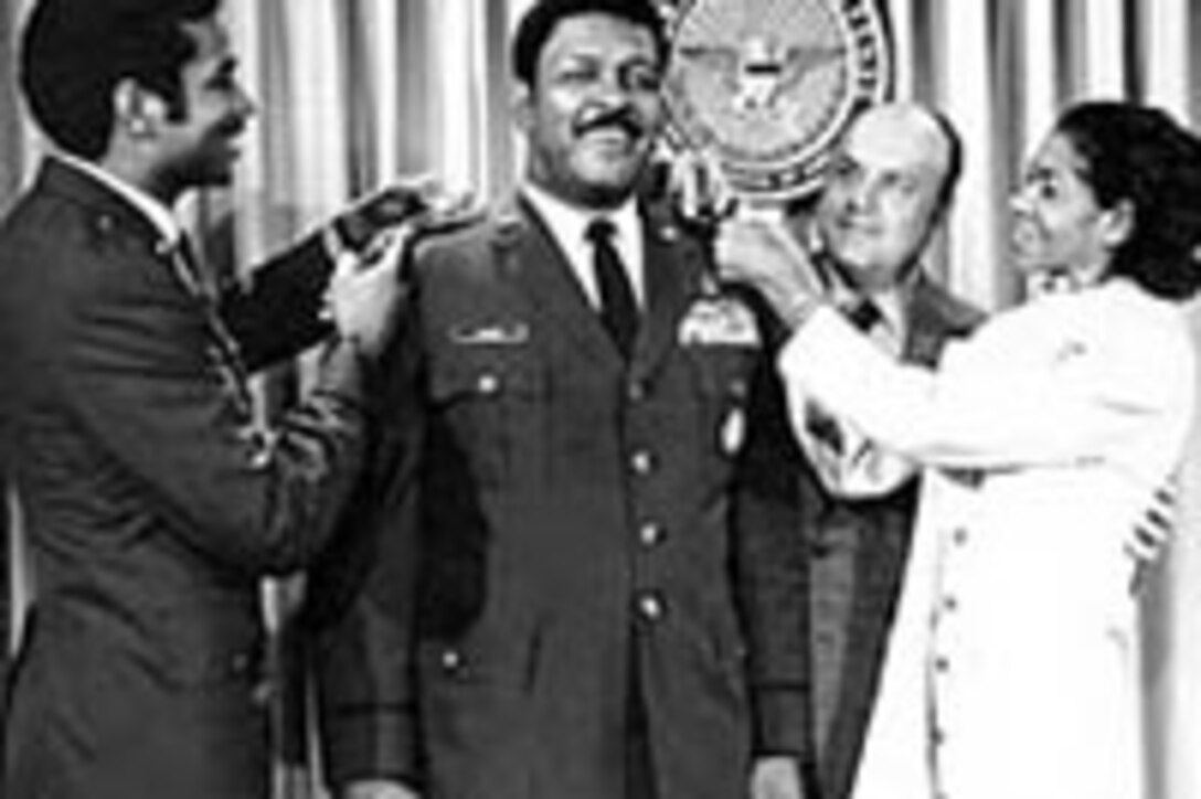 A man in uniform stands as another man and a woman pin something to his shoulders. A fourth person stands watching behind them.