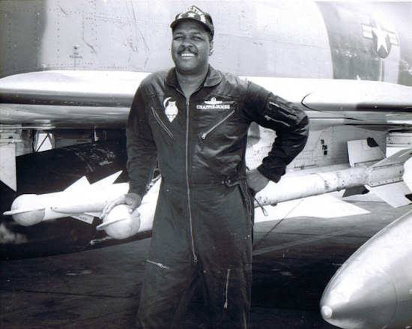 A man in a flight suit stands with his hand on a missile that’s on a rack beside him.