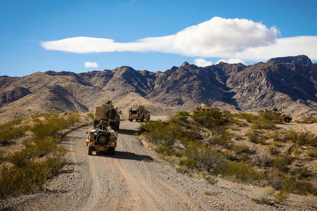 Five military vehicles drive on a rocky road with mountains in the background.