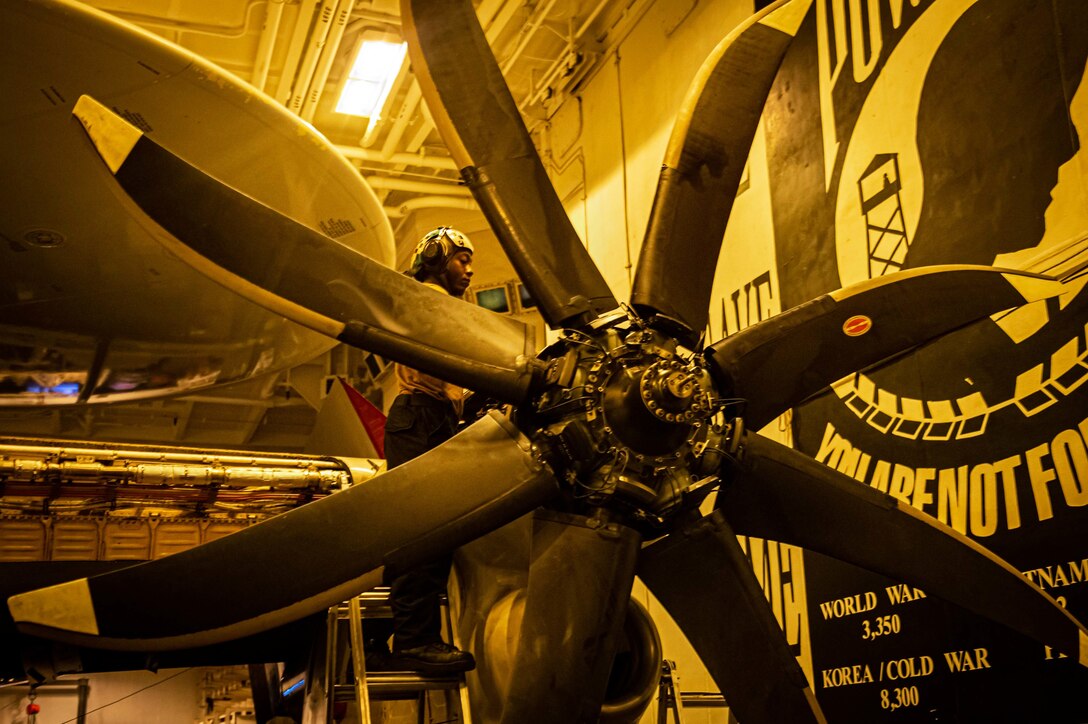 A sailor works on the rotor on an aircraft in a hangar bathed in yellow light.