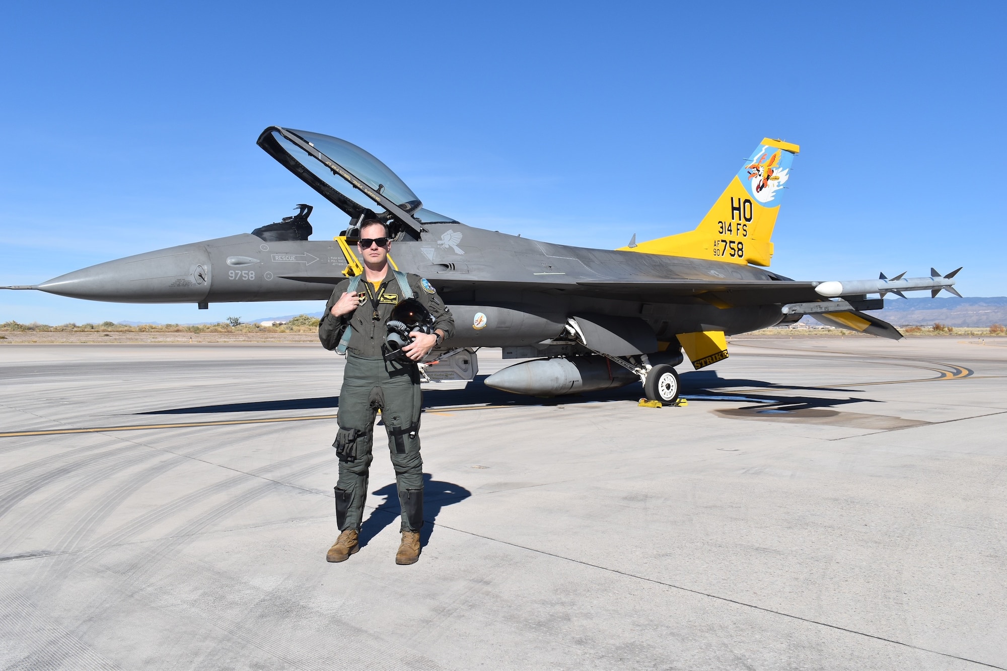 A pilot in a green flight suit poses in front of an F-16 Fighting Falcon with a  yellow tail.