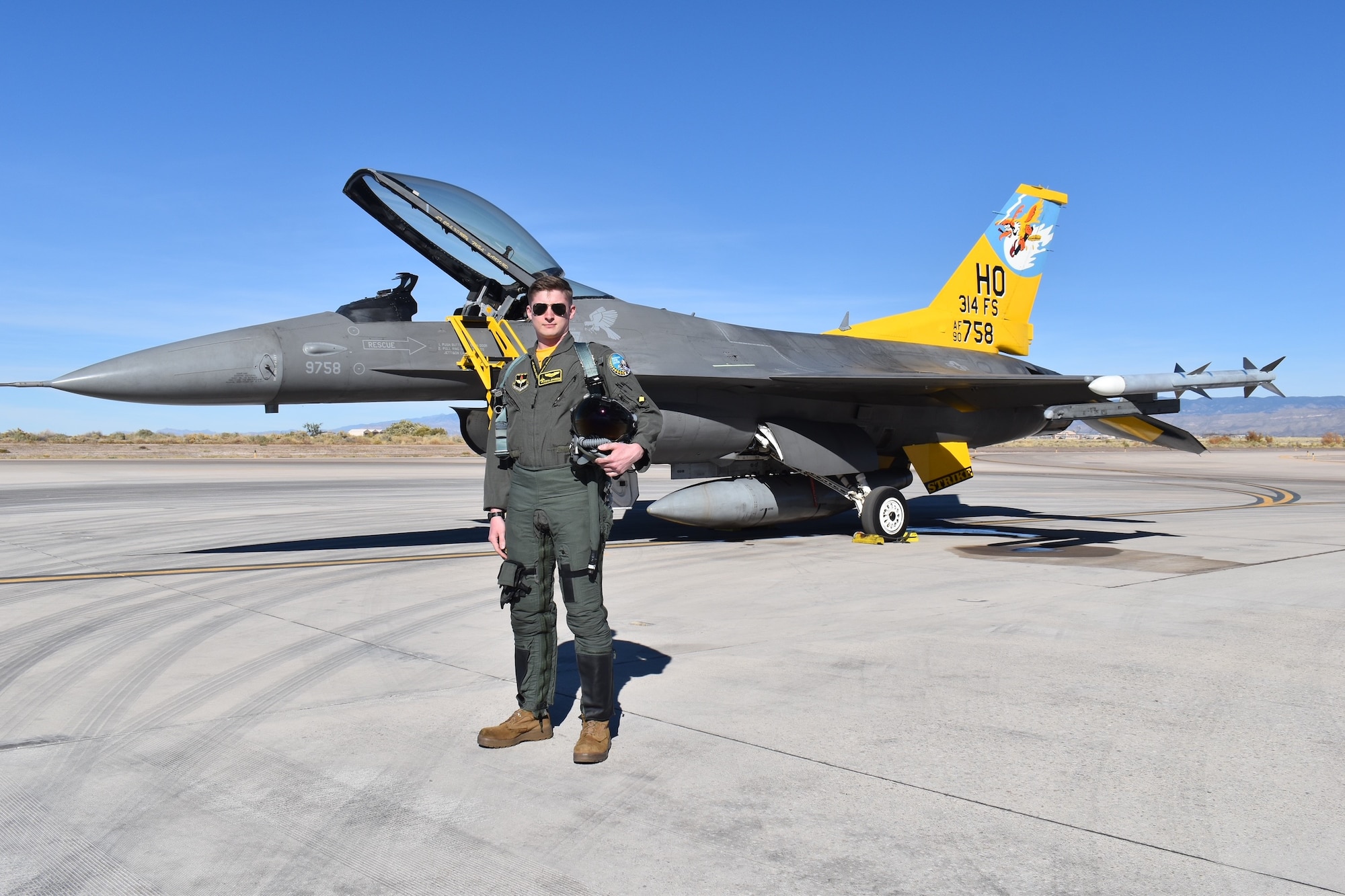 A pilot in a green flight suit poses in front of an F-16 Fighting Falcon with a  yellow tail.