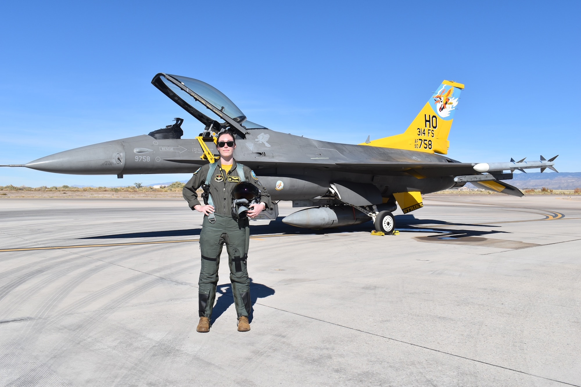 A pilot in a green flight suit poses in front of an F-16 Fighting Falcon with a  yellow tail.