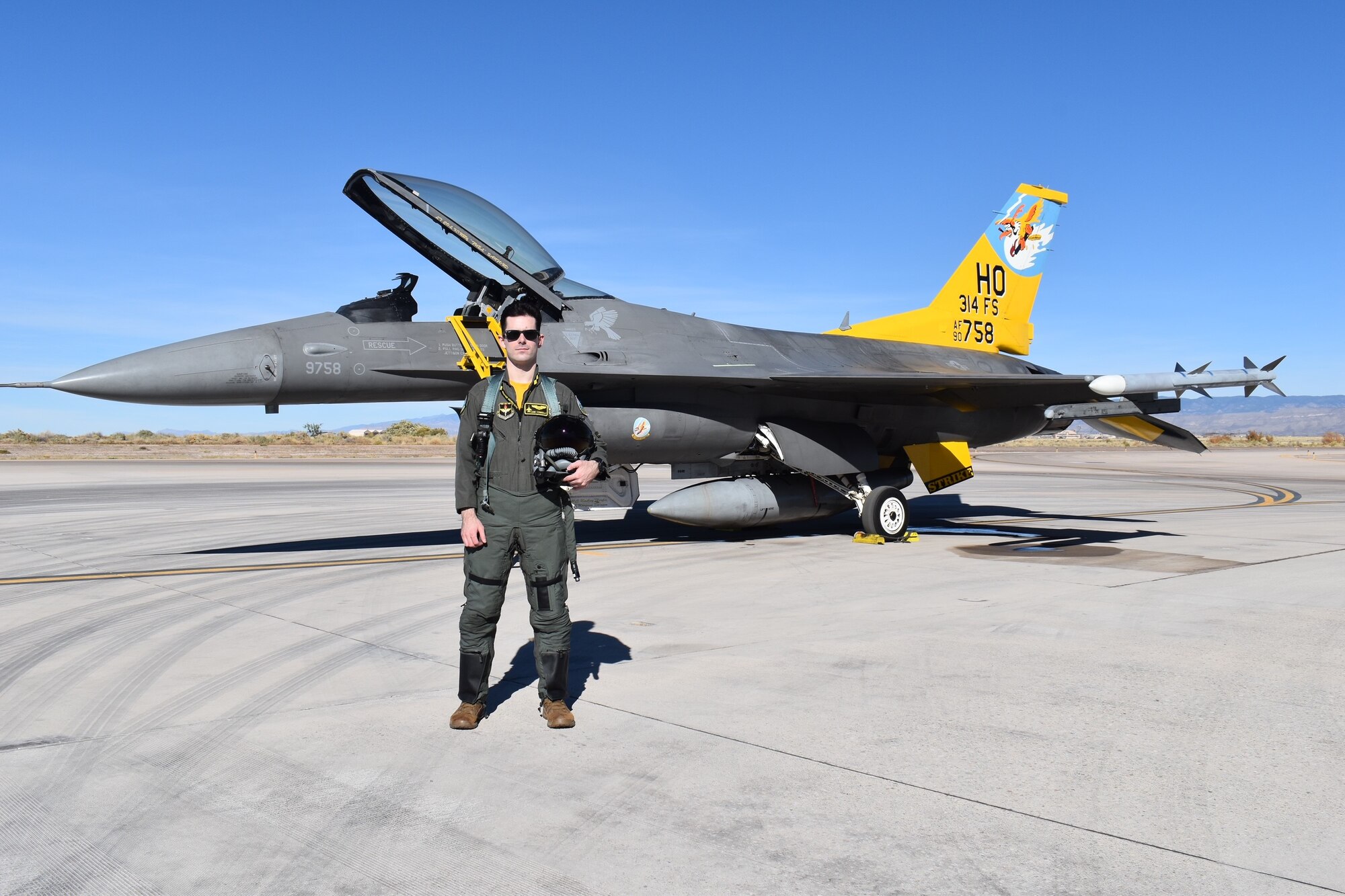 A pilot in a green flight suit poses in front of an F-16 Fighting Falcon with a  yellow tail.