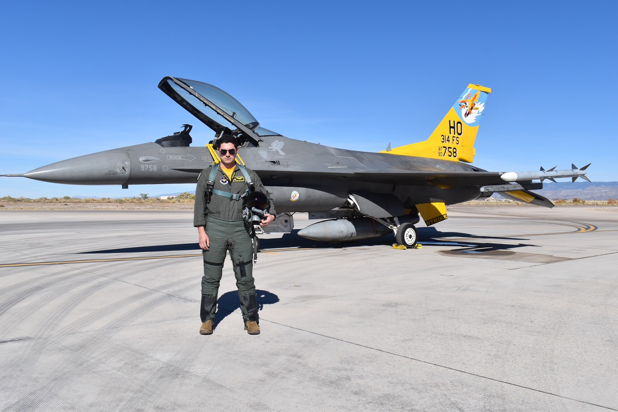 A pilot in a green flight suit poses in front of an F-16 Fighting Falcon with a  yellow tail.