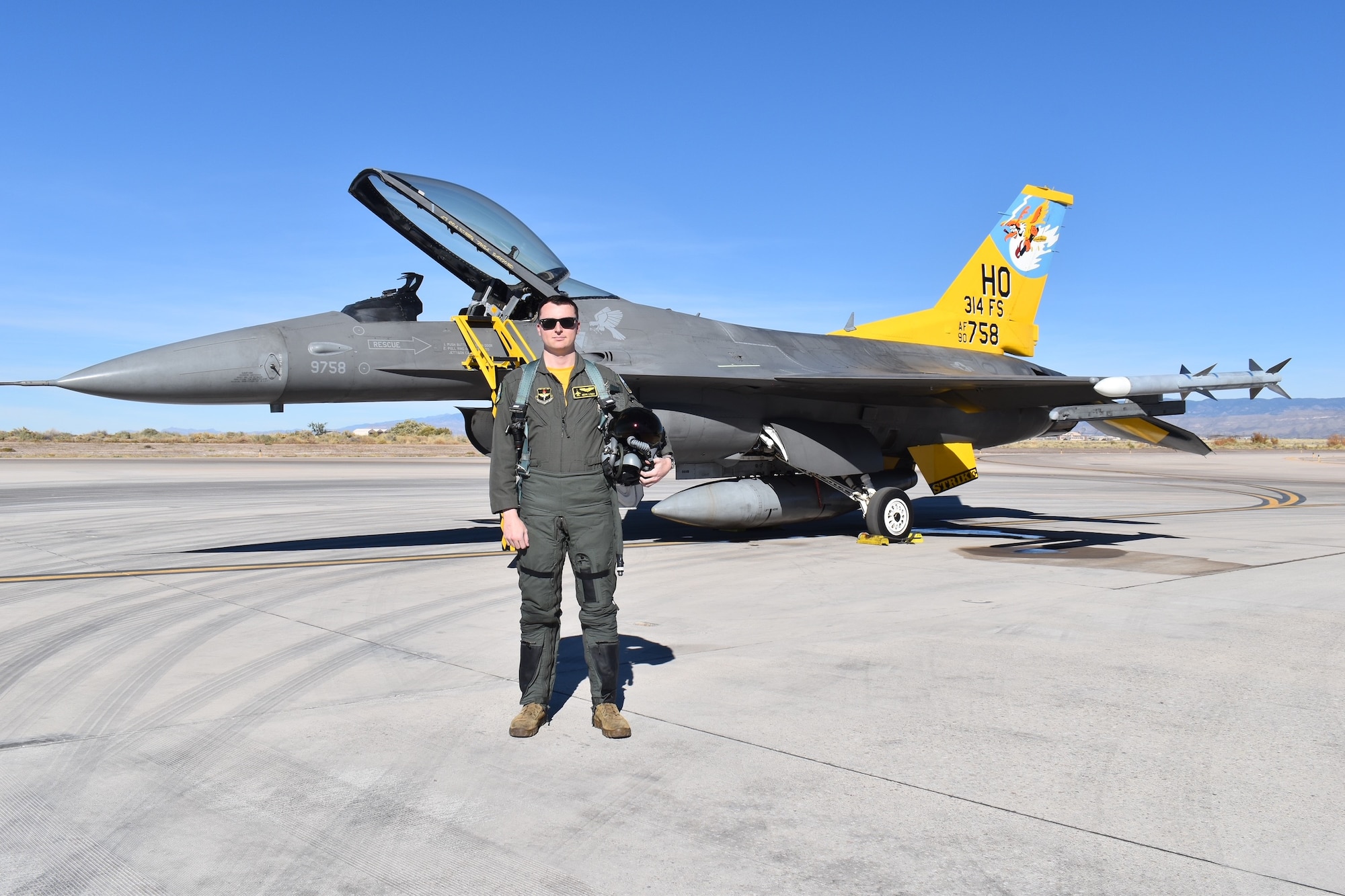 A pilot in a green flight suit poses in front of an F-16 Fighting Falcon with a  yellow tail.