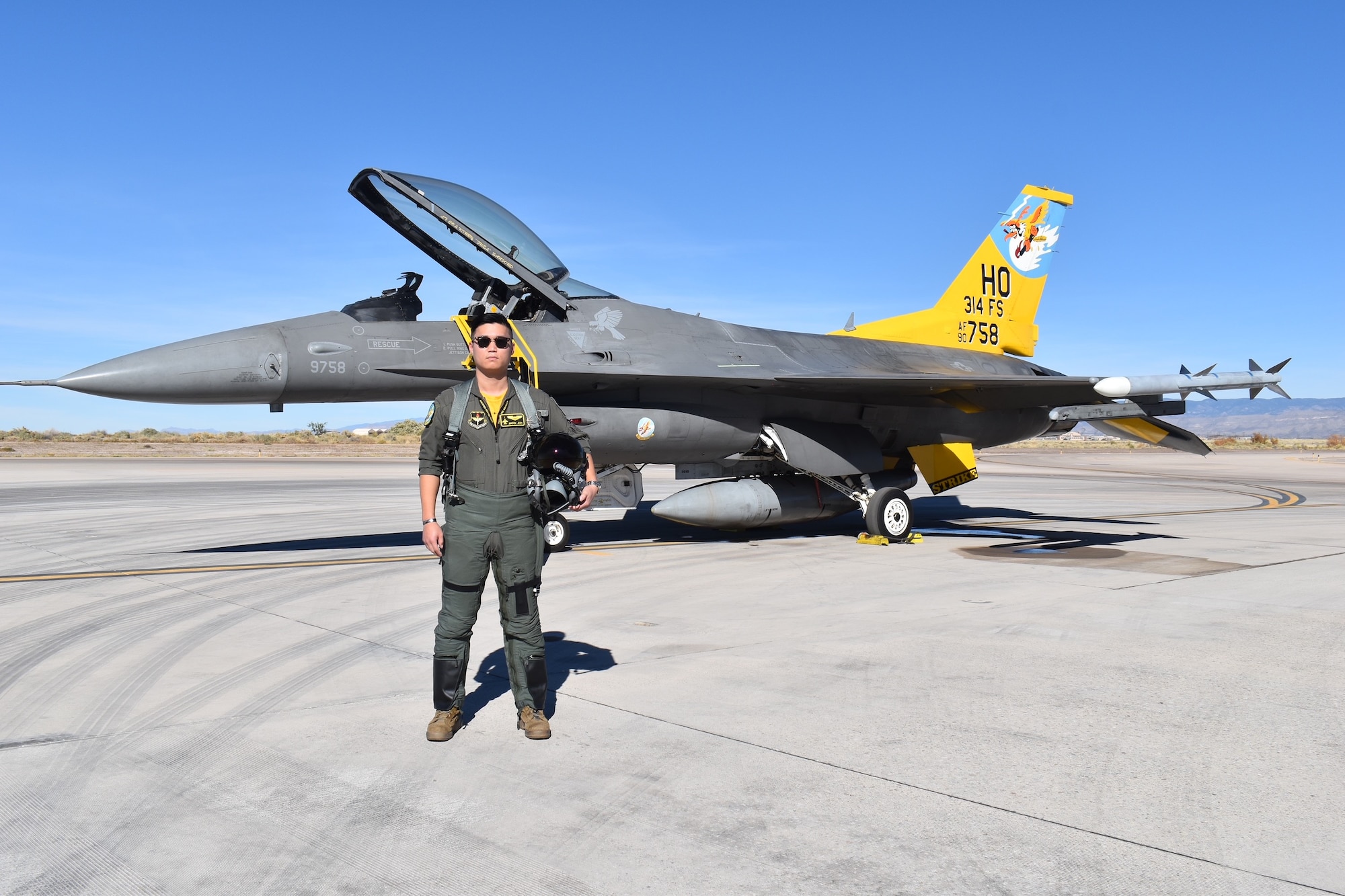 A pilot in a green flight suit poses in front of an F-16 Fighting Falcon with a  yellow tail.