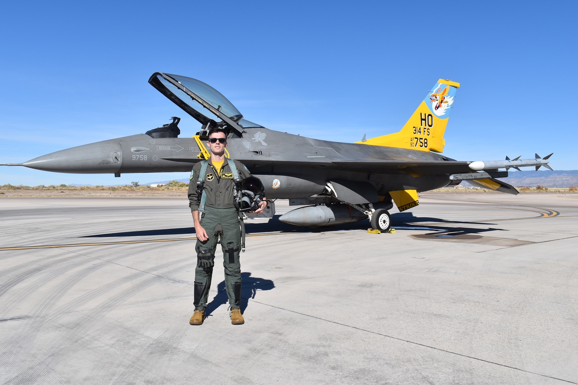 A pilot in a green flight suit poses in front of an F-16 Fighting Falcon with a  yellow tail.