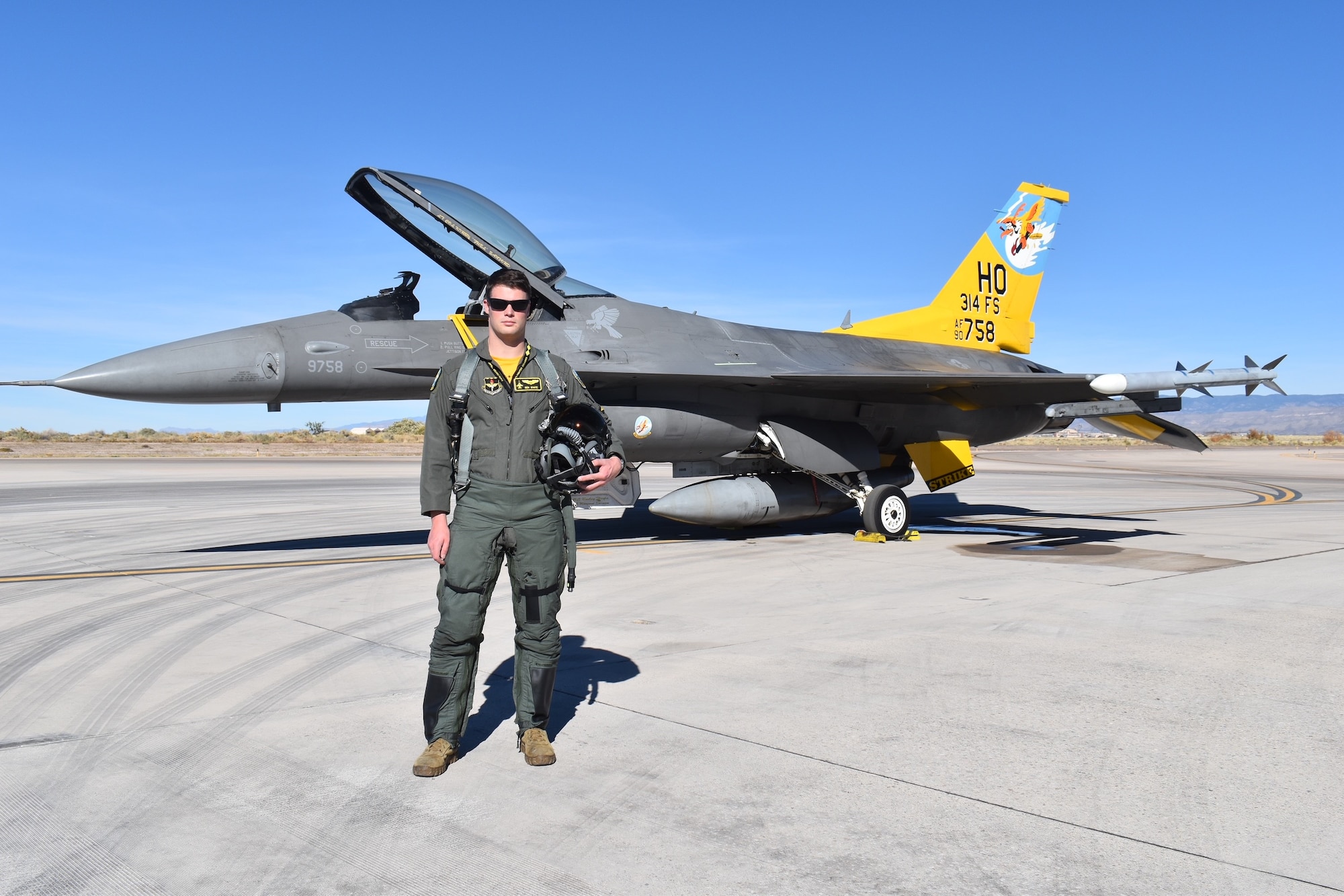 A pilot in a green flight suit poses in front of an F-16 Fighting Falcon with a  yellow tail.