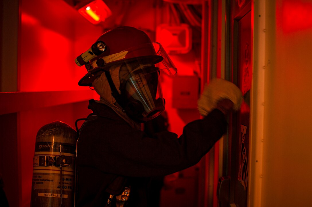 A sailor wearing protective gear touches a door on a ship at sea illuminated by red light.