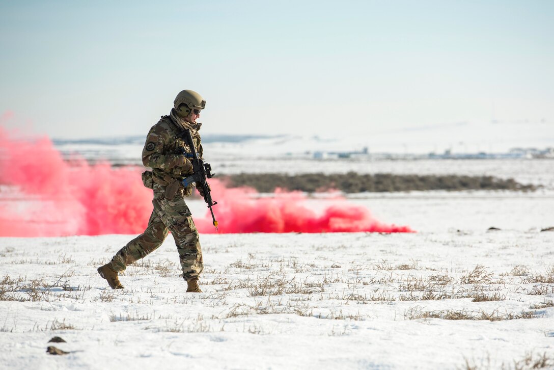 A service member walks through the snow while holding a weapon near a cloud of red smoke.