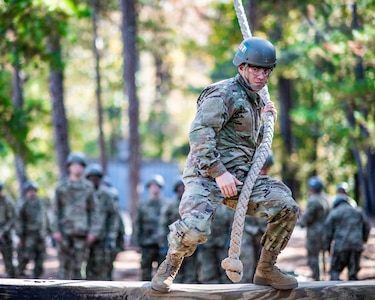 Army trainee in uniform and helmet, holds rope preparing to swing off a raised platform.