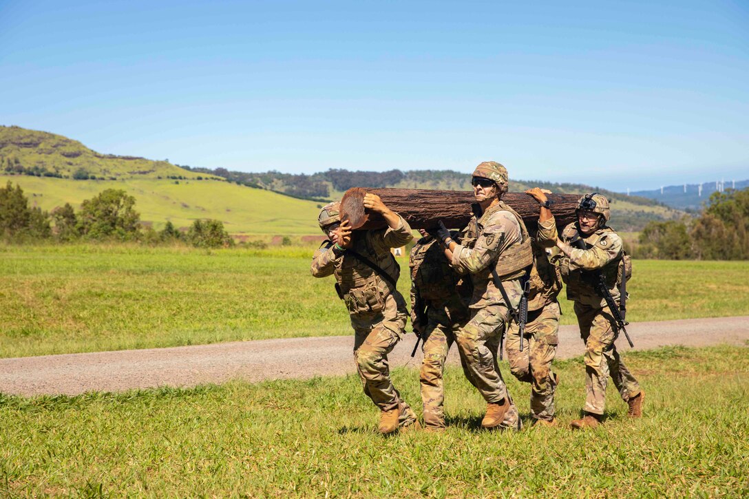 Soldiers lift a log while walking along grass.