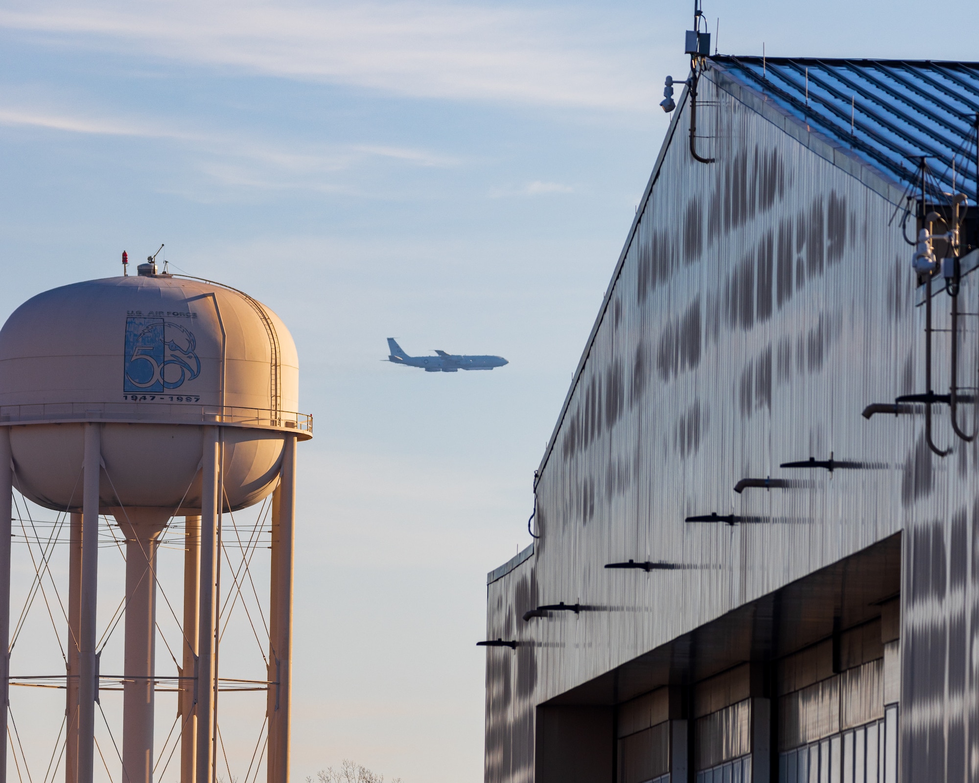 Photo show plane flying in background with a water tower and hangar in foreground.