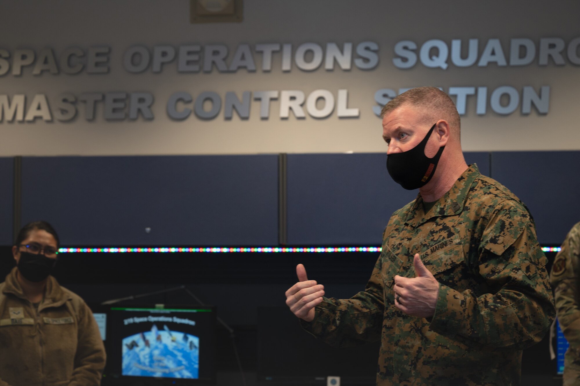 A man stands in a room wearing a cloth facemask with wall lettering that reads "Space Operations Squadron, Master Control Station."