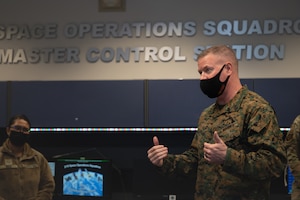 A man stands in a room wearing a cloth facemask with wall lettering that reads "Space Operations Squadron, Master Control Station."