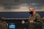 A man stands in a room wearing a cloth facemask with wall lettering that reads "Space Operations Squadron, Master Control Station."