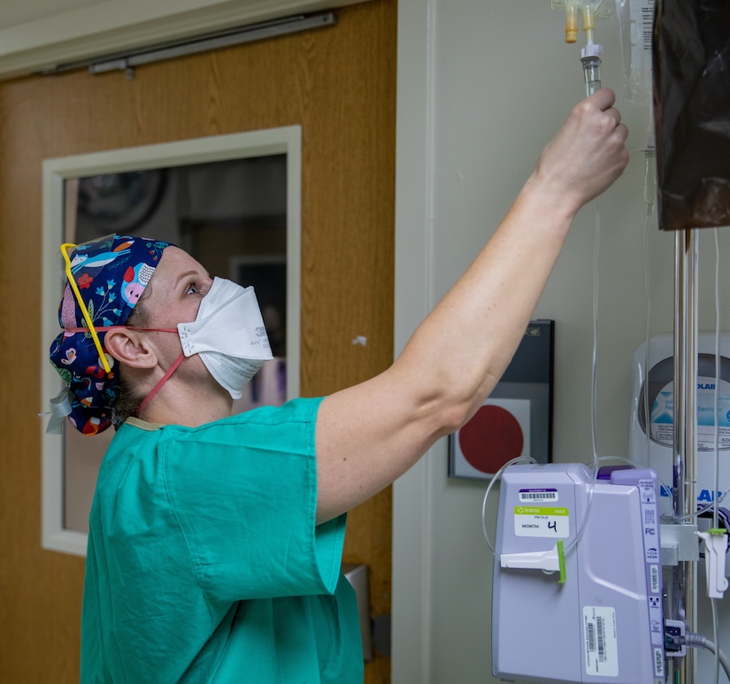 A woman in a medical gown and face mask checks on an IV fluid tube.