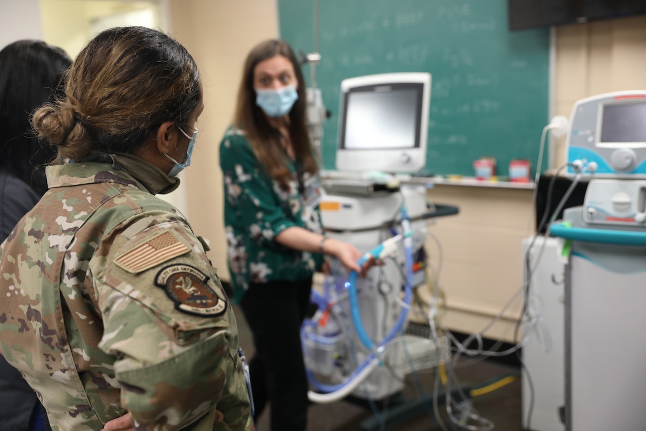 A woman in a face mask holds medical equipment tubes and speaks to two people.