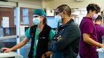 A woman in a face mask holds medical equipment tubes and speaks to two people.