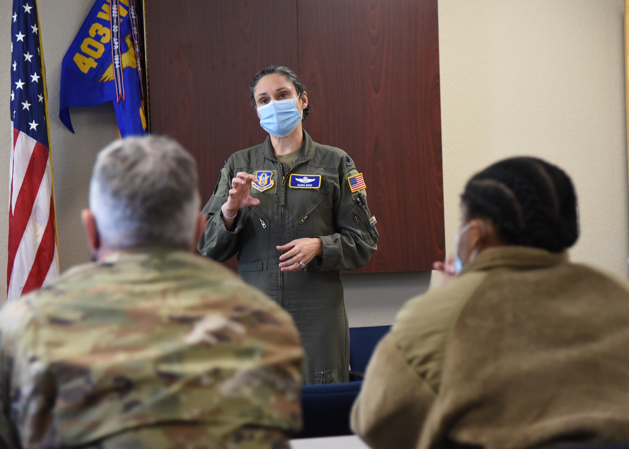 Airman in a flight suit stands in background facing two Airmen sitting down looking forward