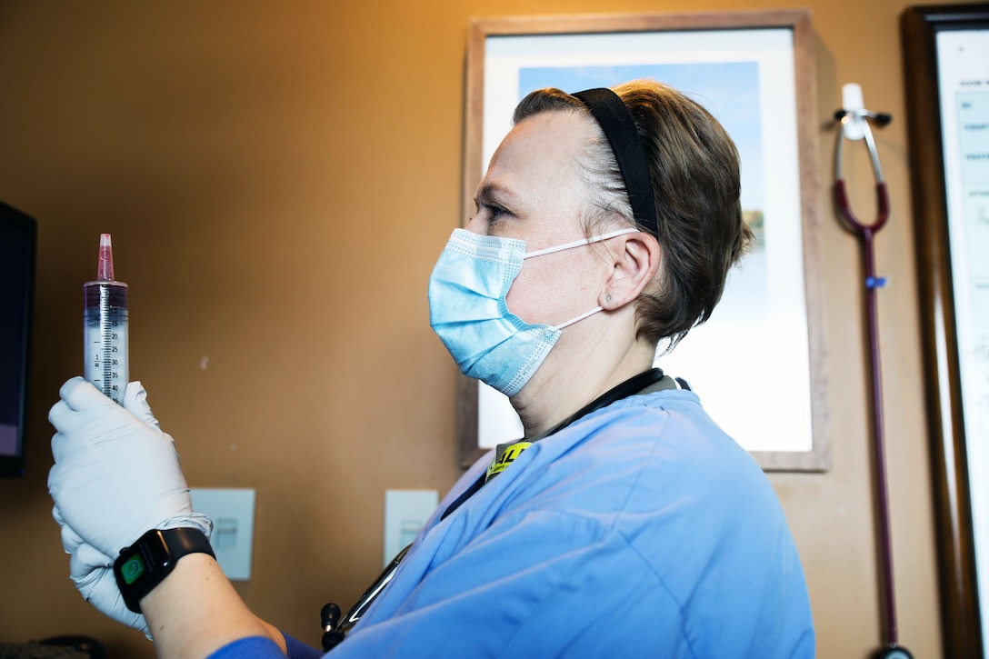 An airman wearing a face mask and gloves holds a syringe to administer medication during a tube feeding.