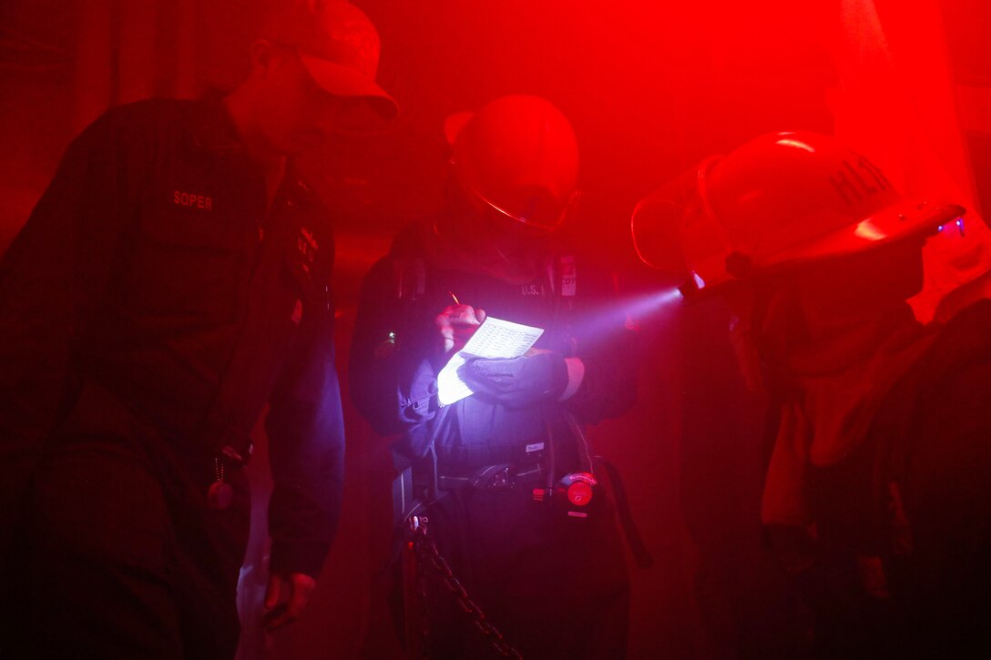 Three sailors use a headlight to look at a piece of paper in a room bathed in red light.