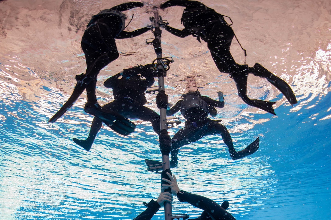 Divers underwater gather around a pole near the surface.