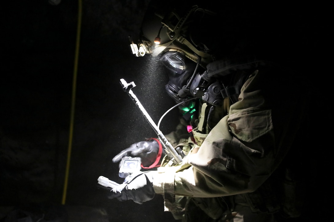 A soldier wearing protective gear holds a small monitor while standing in a dark area.