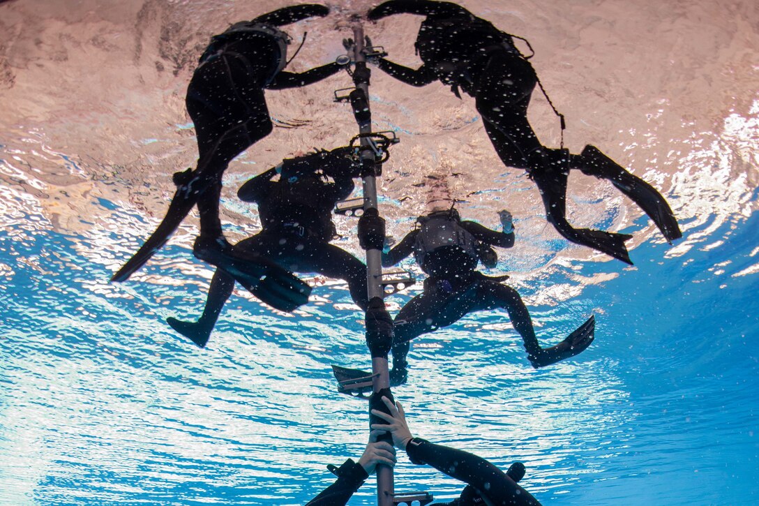 Divers underwater gather around a pole near the surface.