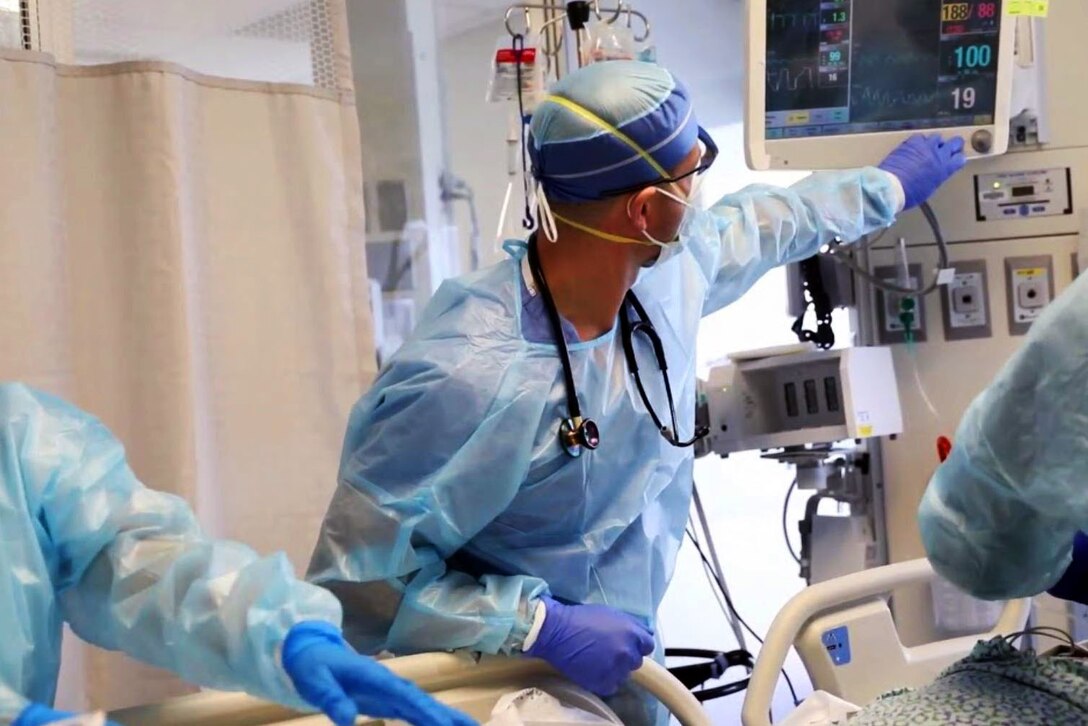 A man wearing a face mask, gloves, surgical gown and cap adjust equipment in a patient’s room.