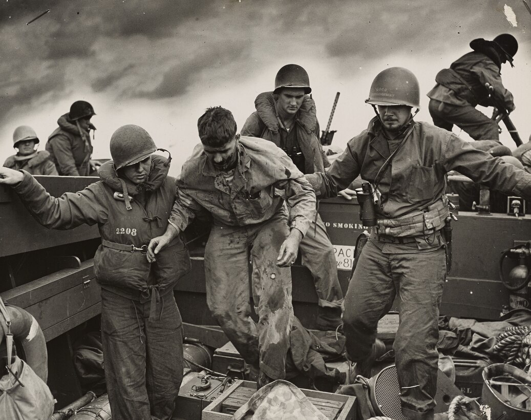 Two men hold the arms of a third man to help him walk as other men move in the background.