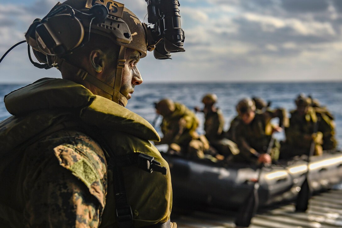 A Marine looks out as fellow Marines exit a ship at sea on a small boat.