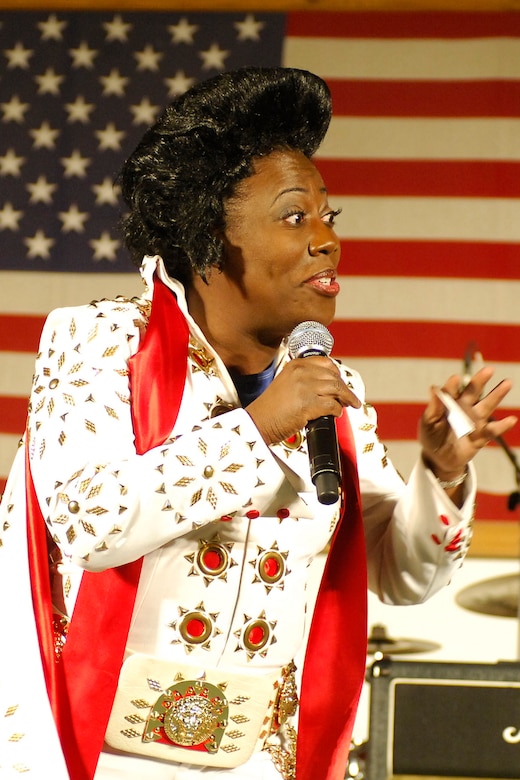 A woman in costume holds a microphone. She is standing in front of a U.S. flag.